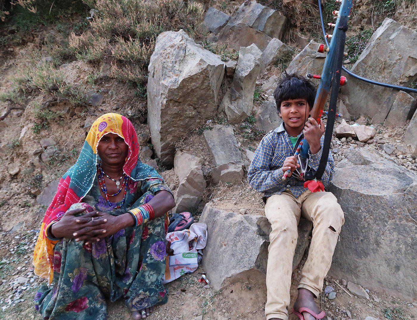 A woman and a young boy sitting amongst rocks. The boy has an instrument called ravanahatha in his hands