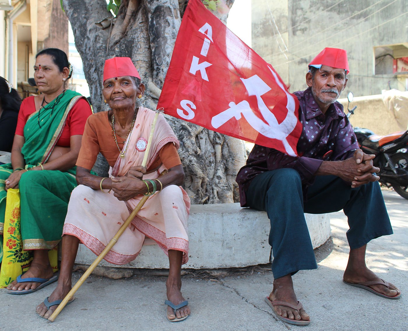 Two women and a man sitting under a tree. One of the women is holding the All India Kisan Sabha flag.