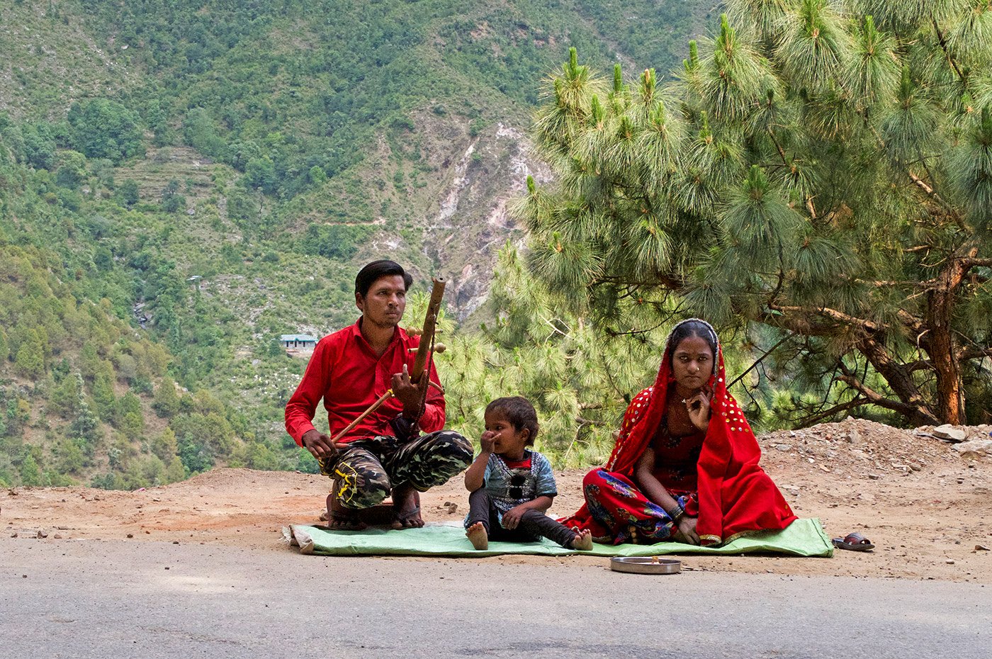 A man and his wife sitting on the side of the road in the moutains. the man is holding an instrument called ravanahatha in his hands