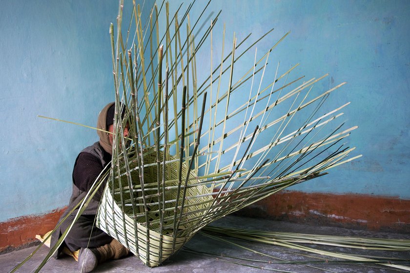 A man is sitting on the floor inside his house and weaving bamboo strips into a basket