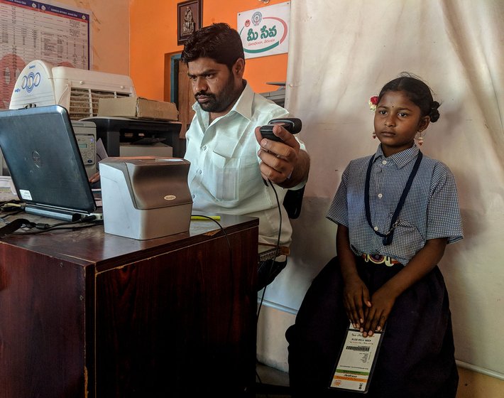 A man sitting at a desk in an office taking a photograph of a young girl in a school uniform. She is holding her Aadhaar card.