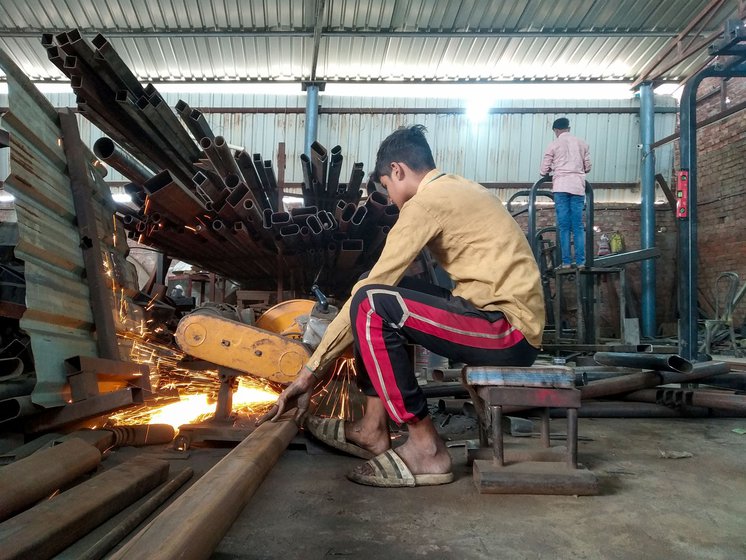 Asif pushes the iron pipe along the empty floor on his left to place it on the cutting machine; he cuts (right) the 15 feet long iron pipe that will go into making the 8 station multi-gym