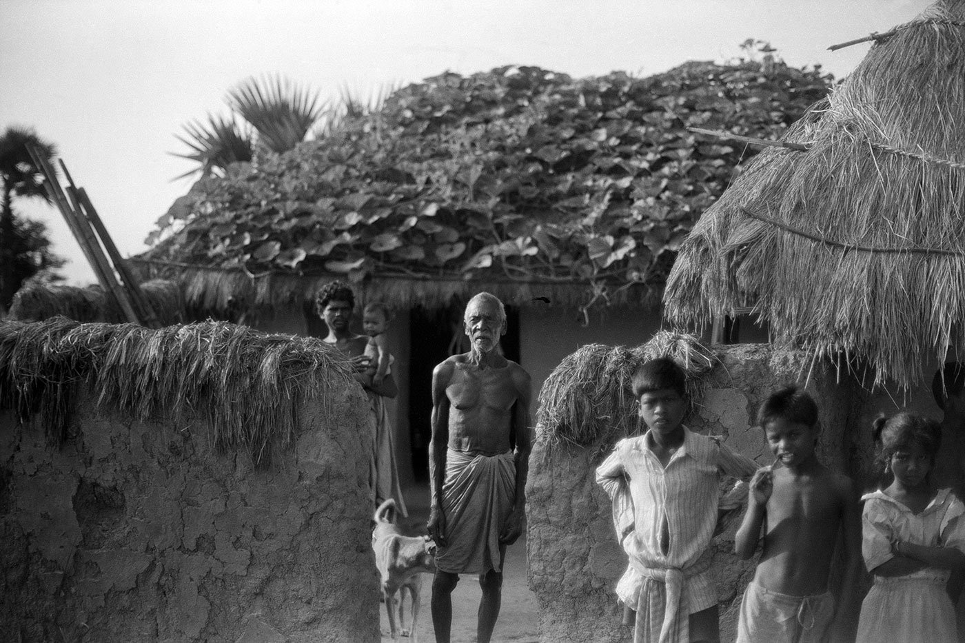 A man standing in front of his hut with three small boys. He has a garden on the roof of his hut.