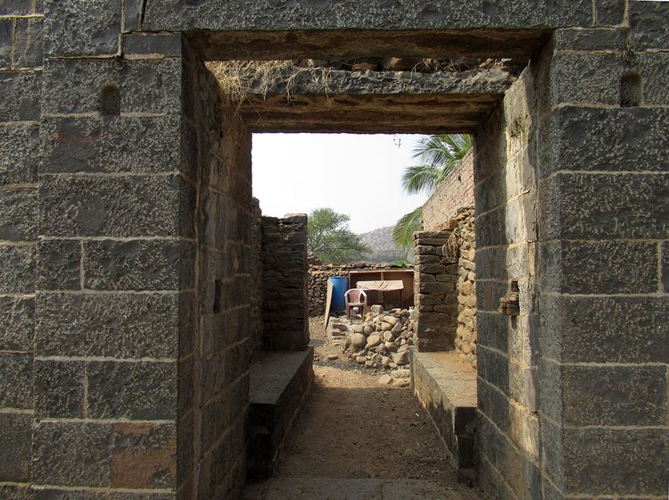 An open doorway made of stone leading into a long passage. There is an empty chair and a pile of stones at the end of the passage