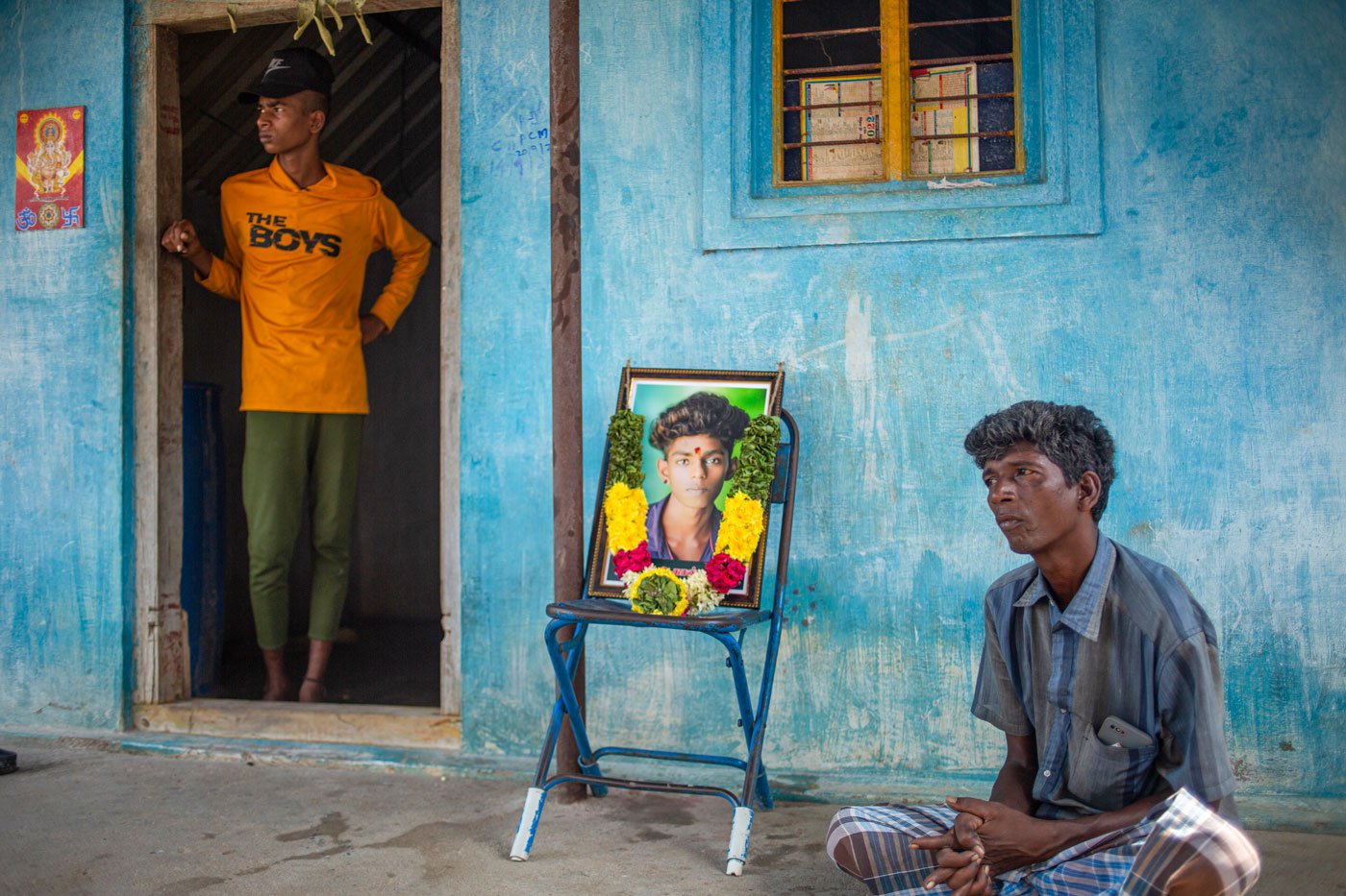 The photograph of another deceased, 19-year-old Akash, is garlanded and placed on a chair in front of the house. His father, M. Raja (right)
