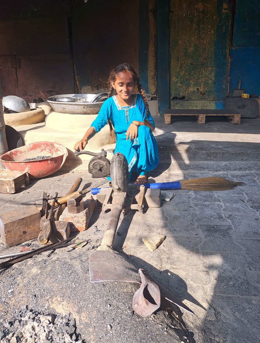 Nine-year-old Chidiya uses a hand-operated fan to blow the ashes away from the unlit bhatti . The family earn much less these days than they did just a few years ago – even though they work in the middle of a busy market, sales have been slow since the pandemic